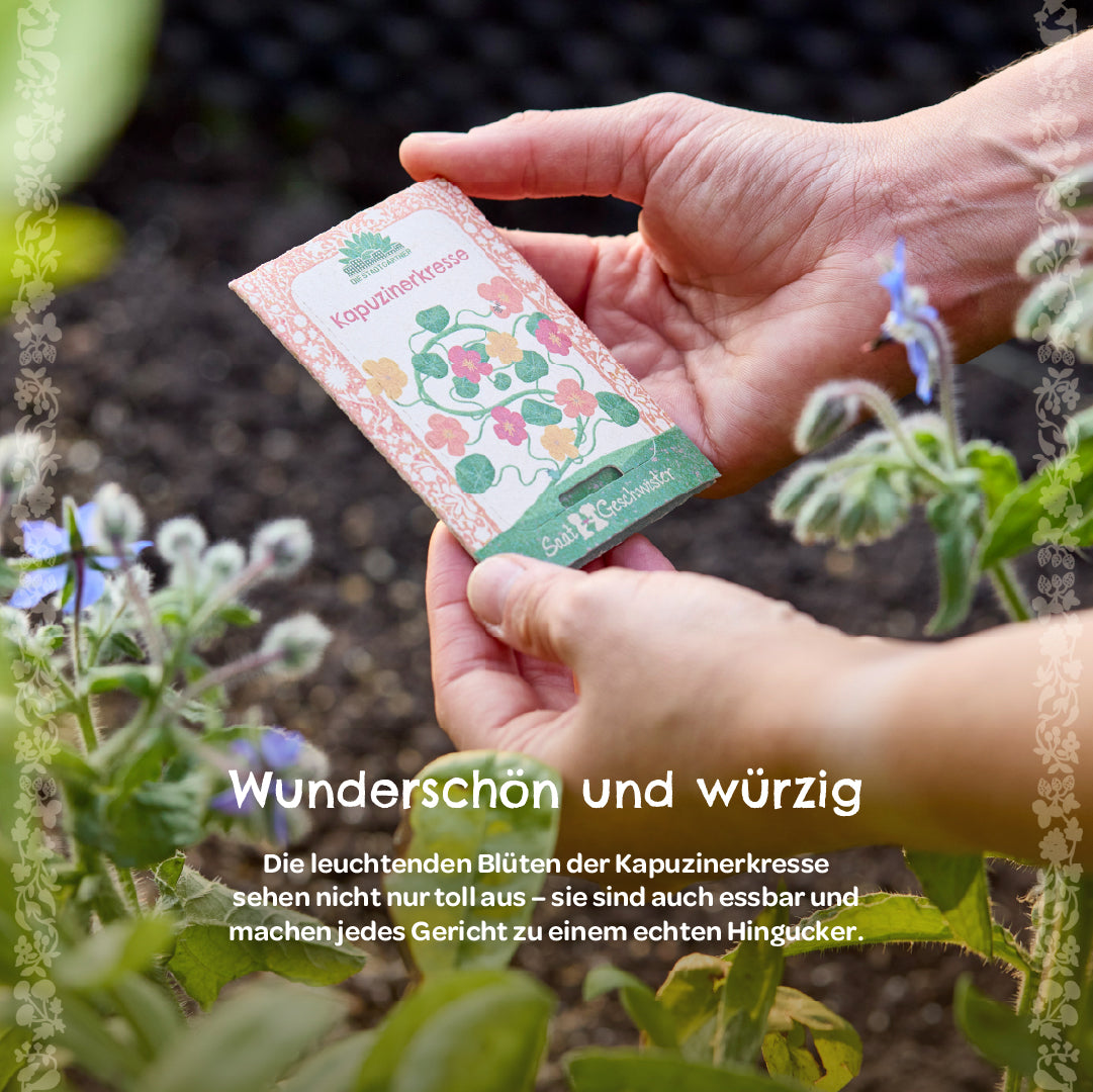 Hände halten ein Samentütchen mit der Aufschrift „Kapuzinerkresse“ im Garten, daneben blühende Pflanzen.