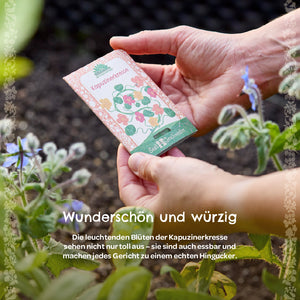 Hände halten ein Samentütchen mit der Aufschrift „Kapuzinerkresse“ im Garten, daneben blühende Pflanzen.