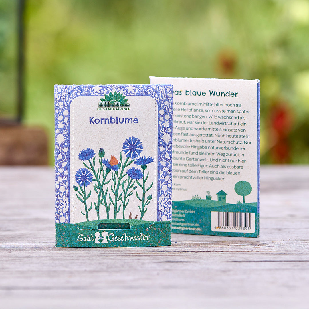 Zwei Samentütchen für Kornblumen der Marke "Saatgeschwister" auf einem Holztisch, mit floraler Illustration und Produktbeschreibung, im Hintergrund unscharfe Natur.