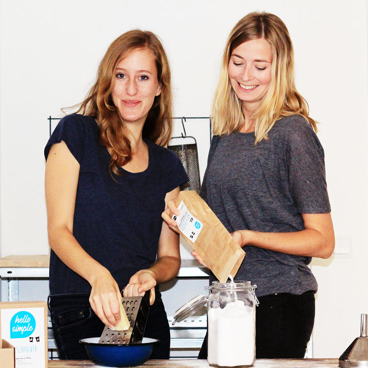 Zwei lächelnde Frauen stehen an einer Küchentheke, wobei eine Käse reibt und die andere eine Papiertüte hält.