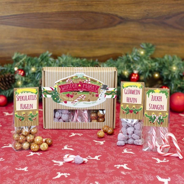 Weihnachtliche Geschenkverpackung mit Spekulatiuskugeln, Glühweinherzen und Zuckerstangen auf einem Tisch mit festlichem Tischtuch.