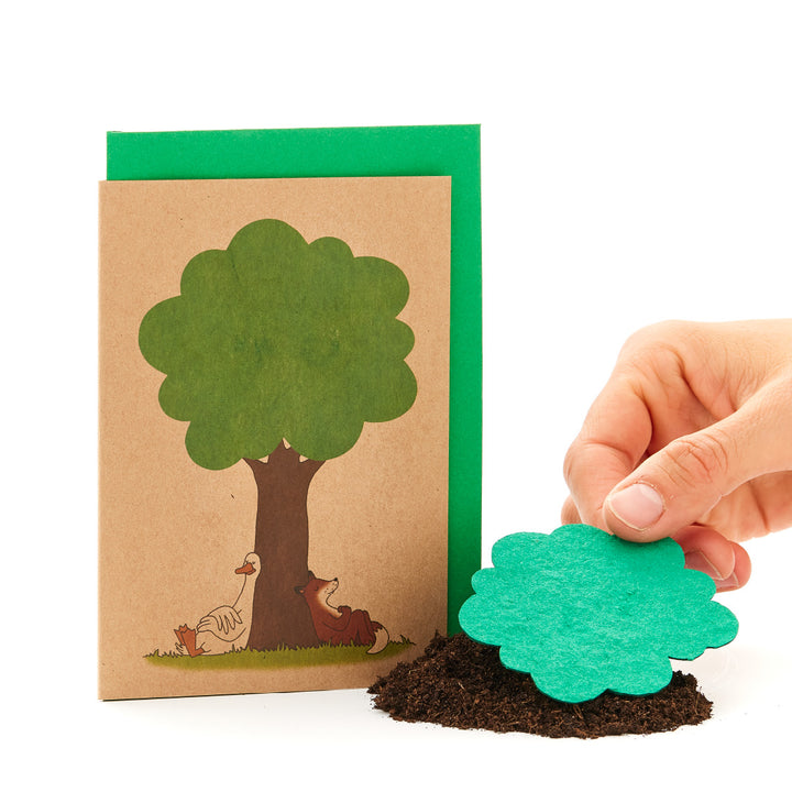 Eine Hand hält eine grüne Papierbaumkrone vor eine Grußkarte mit gezeichneter Baumstamm und Tieren auf weißem Hintergrund.