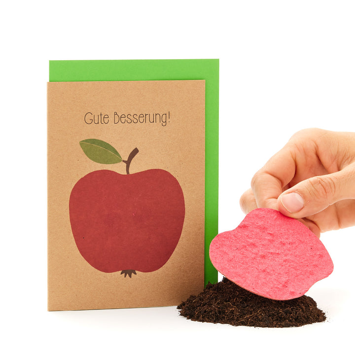 Eine "Gute Besserung"-Karte mit einem Apfel drauf neben einer Hand, die eine herzförmige Samenkarte in Erde pflanzt.