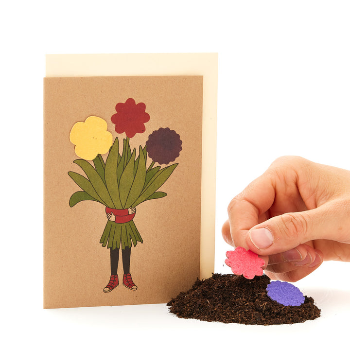 Hand legt eine Papierblume auf einen Haufen Erde neben einer Grußkarte mit Blumenstrauß-Design."