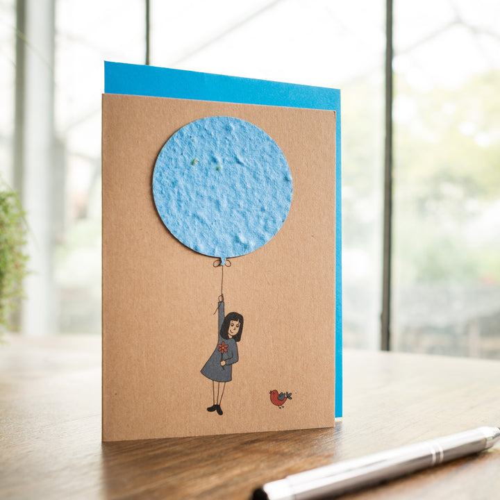 Handgemachte Grußkarte mit aufgezeichneter Figur, die an einem Ballon schwebt, und einem Vogel auf Holztisch.
