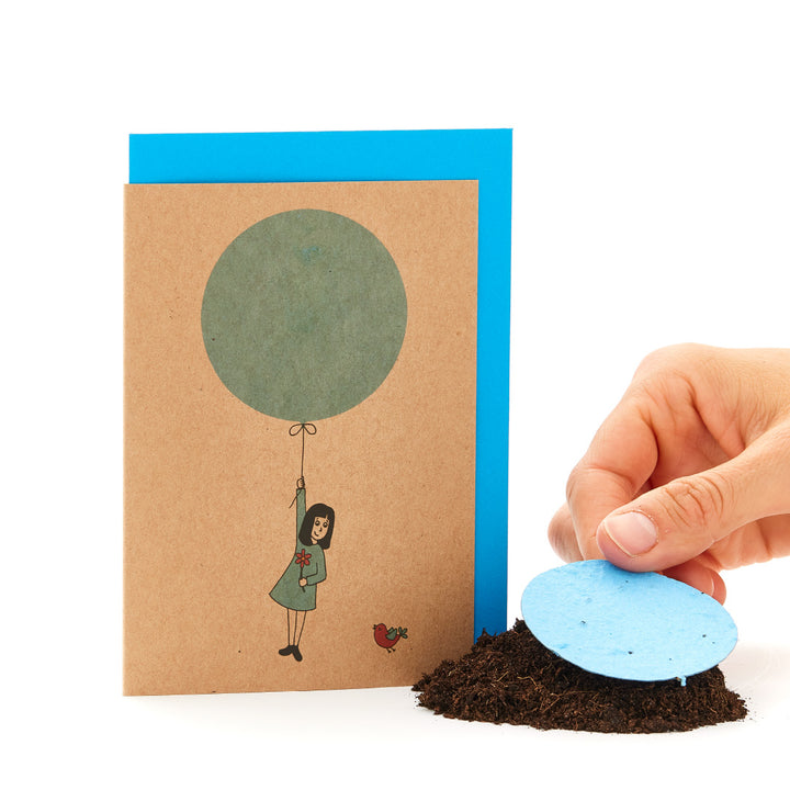 Eine Grußkarte mit einer gezeichneten Figur, die an einem Ballon hängt, und eine Hand, die ein blaues rundes Samenpapier auf Erde legt.
