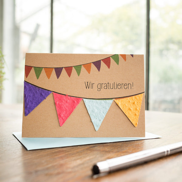 Glückwunschkarte mit der Aufschrift "Wir gratulieren!" und bunten Wimpeln auf Holztisch mit Stift und Briefumschlag.
