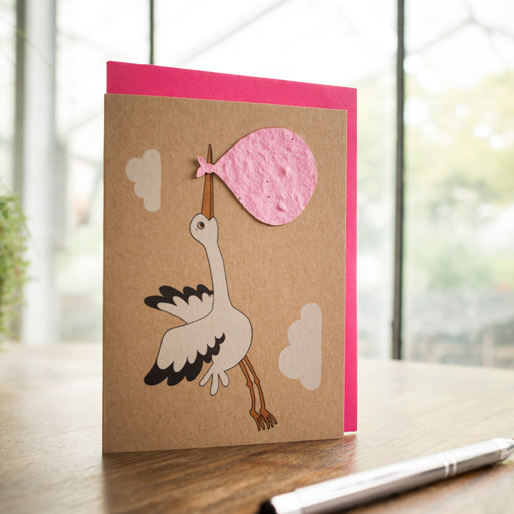 Glückwunschkarte mit Storchmotiv, der ein rosa Bündel trägt, auf einem Holztisch mit Stift daneben.