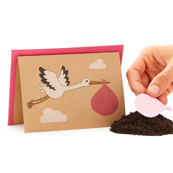 Storch auf einer Grußkarte, der ein Babybündel trägt, neben einer Hand, die ein rosa herzförmiges Objekt auf einem Haufen Erde hält.