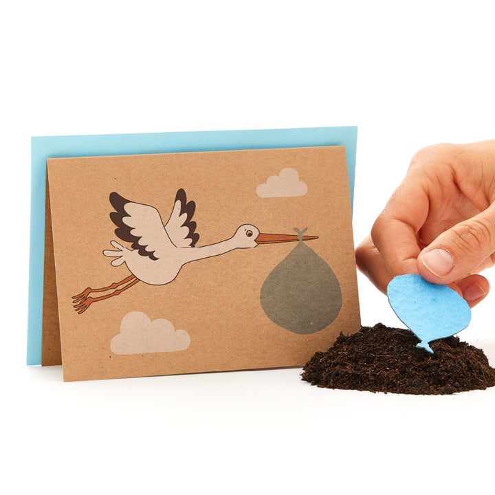 Grußkarte mit Storch, der ein Babybündel trägt, neben einer Hand, die einen blauen Herz-förmigen Samen in die Erde pflanzt.