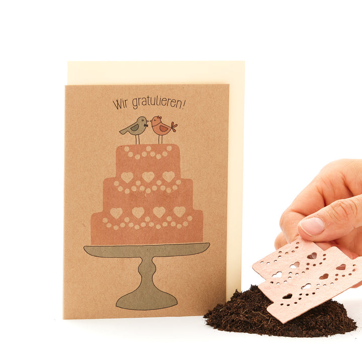 Glückwunschkarte mit einem aufgedruckten Kuchen und zwei Vögeln sowie einer menschlichen Hand, die ein Stück Papier hält.