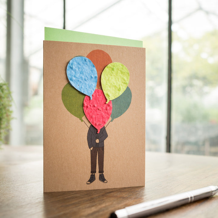 Grußkarte mit Illustration einer Person, deren Kopf aus bunten Papierballons besteht, auf einem Tisch neben einem Stift.