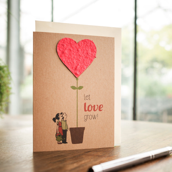 Grusskarte mit einem Paar, das sich küsst, neben einem Topf, aus dem eine Pflanze mit einem roten Herzen wächst, mit dem Text "let love grow!"