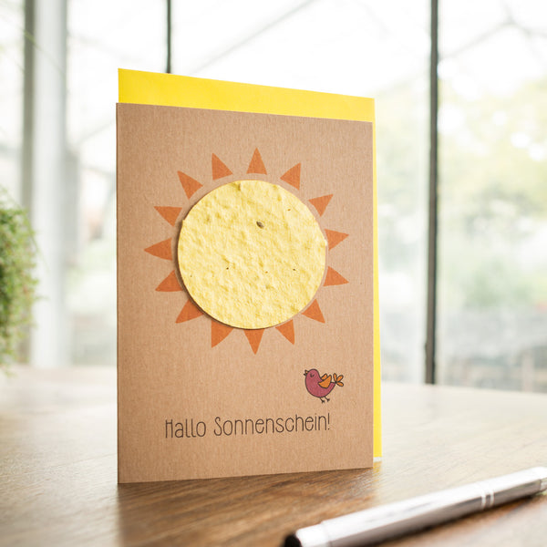 Handgemachte Grußkarte mit Sonne und der Aufschrift "Hallo Sonnenschein!" samt kleinem Vogel auf einem Holztisch.