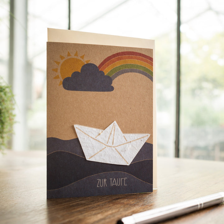 Glückwunschkarte zur Taufe mit Papierboot und Regenbogen auf Holztisch.