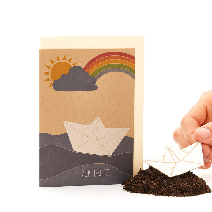 Glückwunschkarte zur Taufe mit Papierboot-Motiv, Regenbogen und Sonne, daneben ein echtes Papierboot auf einem Erdhügel, gehalten von einer Hand.