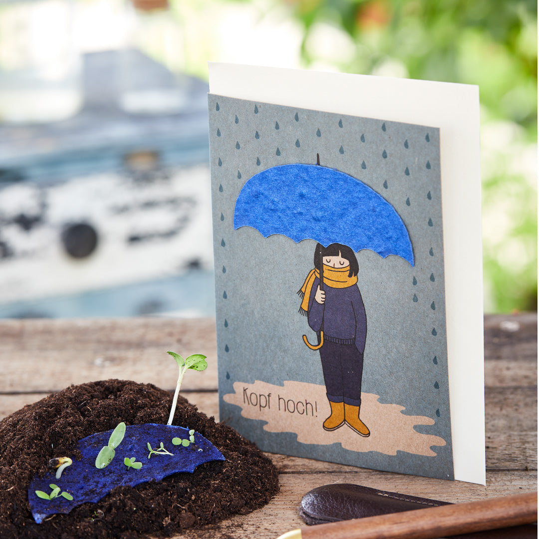 Grußkarte mit Zeichnung einer Person unter einem blauen Regenschirm und der Aufschrift "Kopf hoch!", neben einer jungen Pflanze auf Erde.