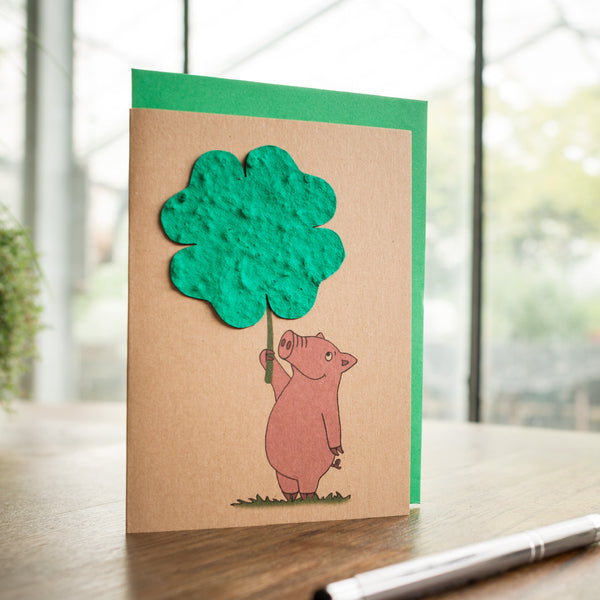 Handgefertigte Grußkarte mit einem gezeichneten Schweinchen, das einen grünen Papierbaum hält, auf einem Holztisch mit Stift.