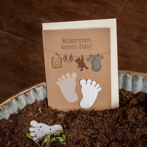 Glückwunschkarte zur Geburt mit der Aufschrift "Willkommen kleines Baby!" und Abbildungen von Babyfüßen, Kleiderbügel mit Strampler und Spielzeug, auf Erde und Holzhintergrund.