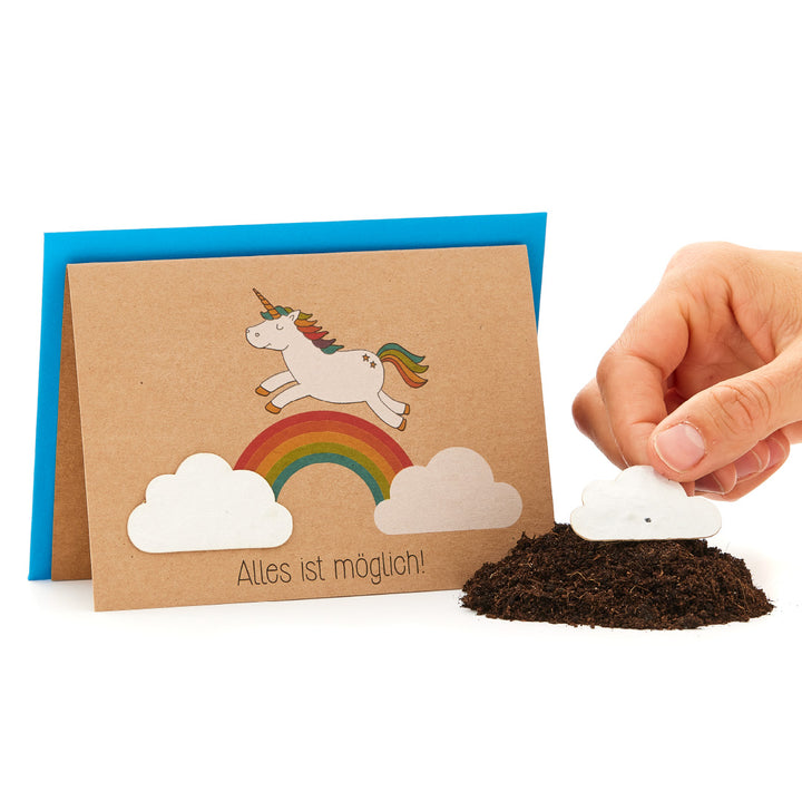 Eine Hand pflanzt eine Papiersaat in Form einer Wolke in Erde, daneben eine Grußkarte mit einem Einhorn auf einem Regenbogen und dem Spruch "Alles ist möglich!".