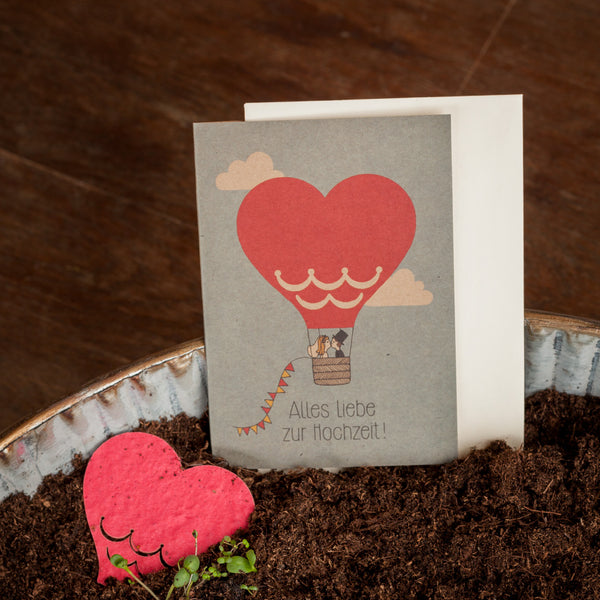 Glückwunschkarte zur Hochzeit mit einem Herzen als Heißluftballon und der Aufschrift "Alles Liebe zur Hochzeit!", platziert neben einem herzförmigen Gegenstand und Pflanzenerde.