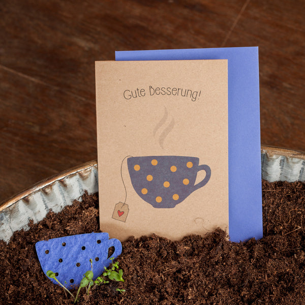 Genesungskarte mit der Aufschrift "Gute Besserung!" und einer illustrierten Tasse auf der Vorderseite, neben einem blauen Pappbecher-Topper in Erde.