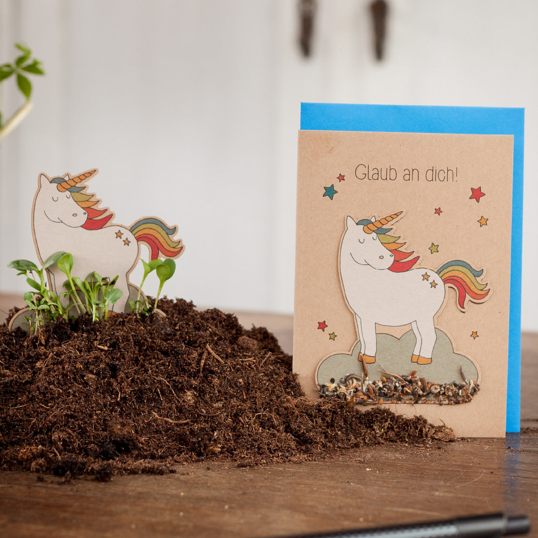 Ein Grußkarte mit einem Einhorn und dem Text "Glaub an dich!" neben einem kleinen Einhorn-Ausschnitt, umgeben von Erde.