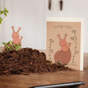 Grußkarte mit Schneckenmotiv und der Aufschrift "blumige Grüße" neben einem Haufen Erde auf einem Holztisch.