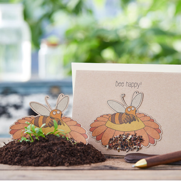 Bastelarbeit mit Bienenmotiv und der Aufschrift "Bee happy!" auf einem Tisch mit Erde und Pflanzen im Hintergrund.