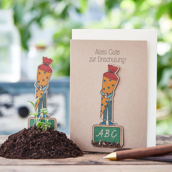 Glückwunschkarte zur Einschulung mit Karotten-Figur und ABC-Tafel auf Holztisch, umgeben von Pflanzen und Erde.