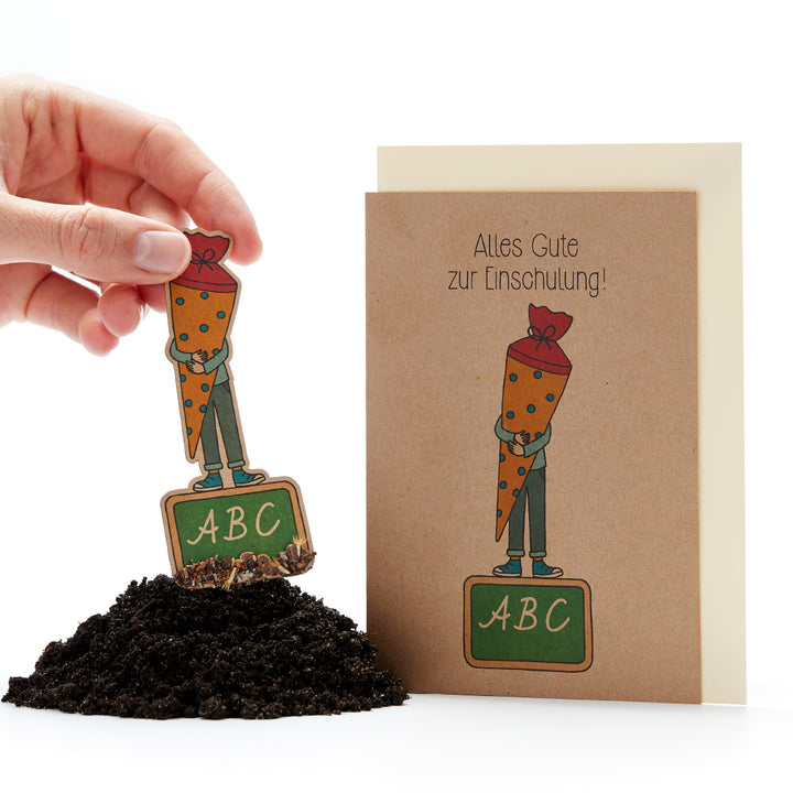 Hand hält eine Karottenfigur mit Schulmütze und ABC-Tafel, daneben eine Glückwunschkarte zur Einschulung.