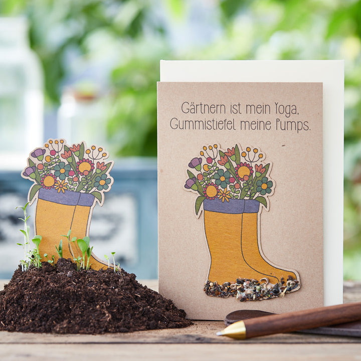 Grußkarte mit dem Spruch "Gärtnern ist mein Yoga, Gummistiefel meine Pumps" und illustrierten Gummistiefeln gefüllt mit Blumen vor einem Hintergrund aus Gartenutensilien.