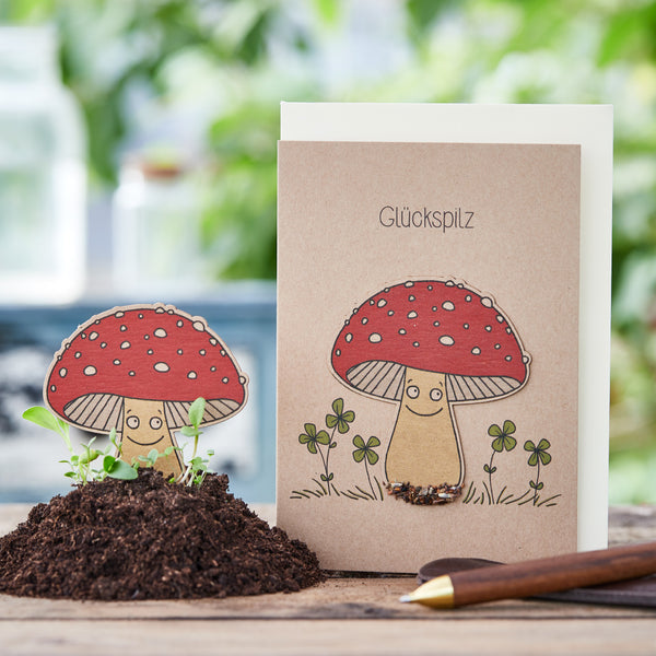 Glückwunschkarte mit zeichnerischen Pilzfiguren und dem Wort 'Glückspilz', neben einem echten Erdhügel und einem Bleistift"