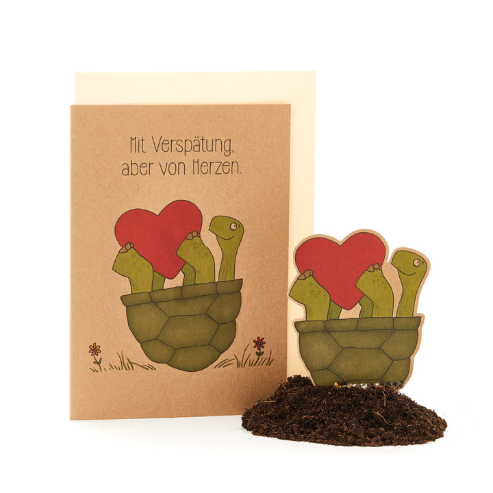 Grüßkarte mit dem Text "Mit Verspätung, aber von Herzen" und einer Illustration von kaktusähnlichen Pflanzen in einem Topf, die herzförmige Blüten tragen.