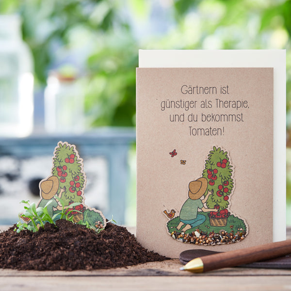 Eine Grußkarte mit dem Spruch "Gärtnern ist günstiger als Therapie, und du bekommst Tomaten!", neben einem Haufen Erde und einer Gartenkelle.