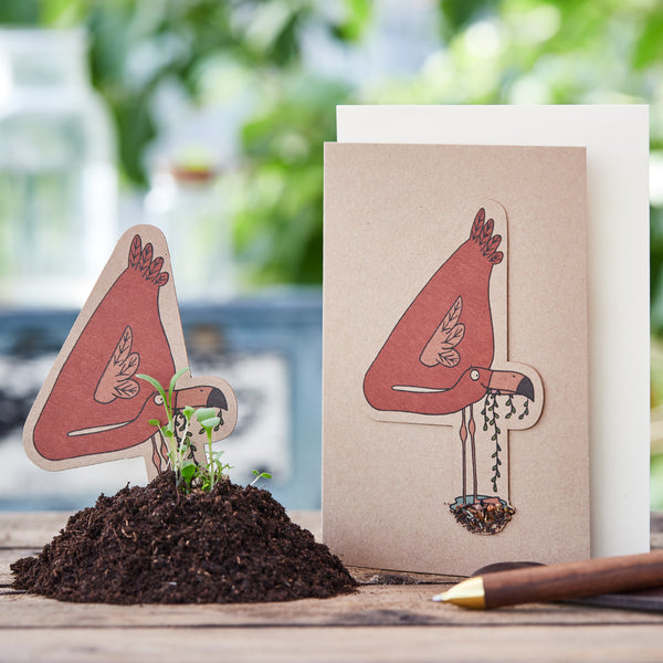 Illustration einer fantasievollen Vogelfigur auf einer Grußkarte neben einer Mound Erde mit kleinen Pflanzen und einem Bleistift, im Hintergrund unscharfes Grün.
