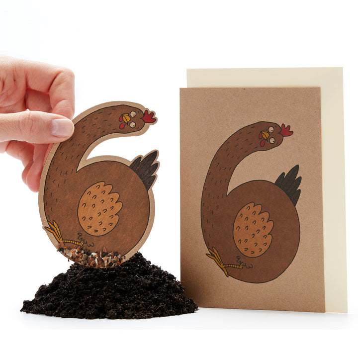 Eine Hand hält eine ausgeschnittene Papierfigur eines Huhns in der Form einer 6, neben einer Karte mit demselben Bild und einem Haufen Erde unten.