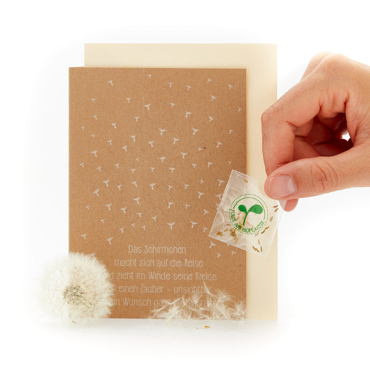 Hand hält eine Grußkarte mit Pusteblumen-Samen und Gedicht neben einer Pusteblume.