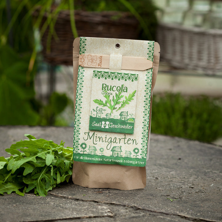 Verpackung für Rucola-Saatgut neben frischen Rucola-Blättern auf Steinuntergrund.