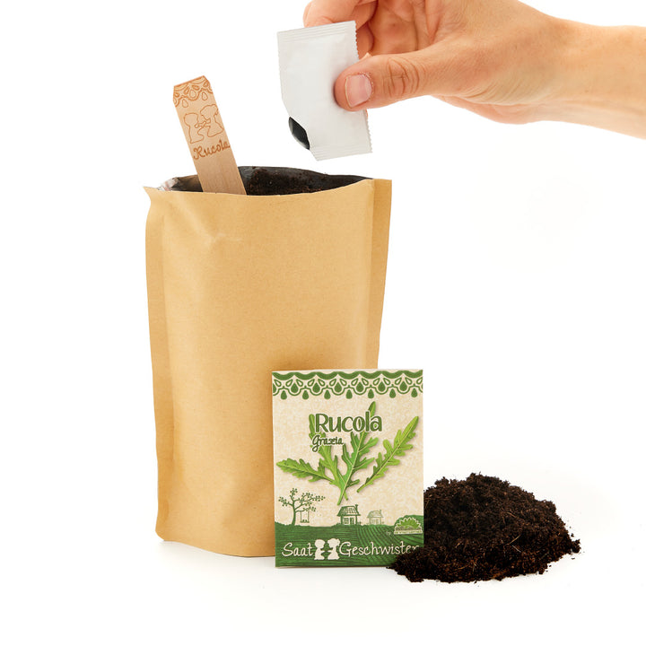 Eine Hand öffnet eine Samenpackung von Rucola neben einer Papiertüte mit Erde und einem Holzschild mit der Aufschrift "Rucola".