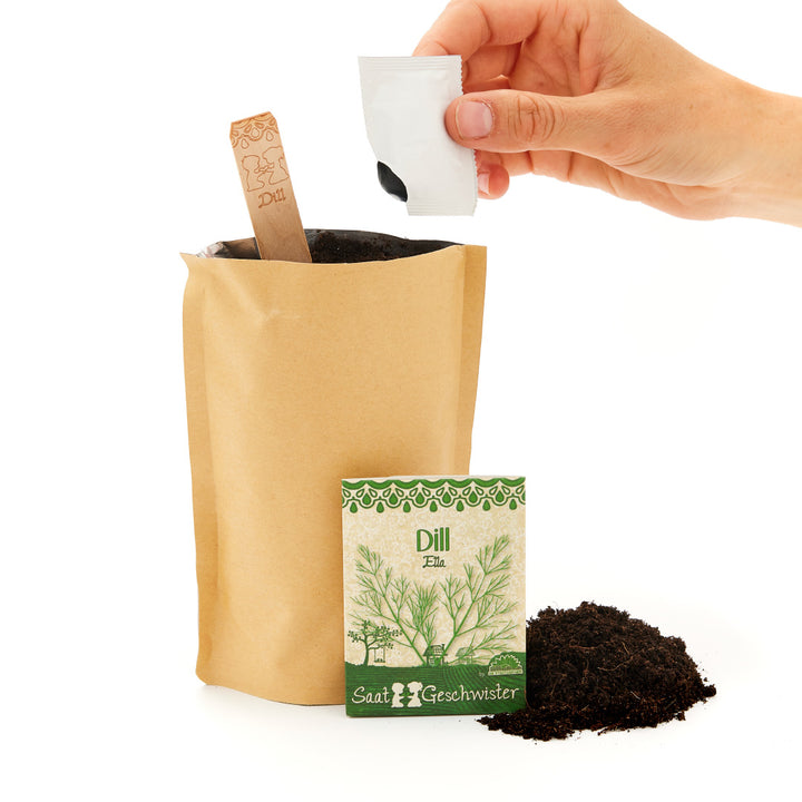 Hand, die Samen in einen Papiertüten-Pflanzbehälter gießt, daneben eine Packung Dillsamen und eine Schaufel Erde.