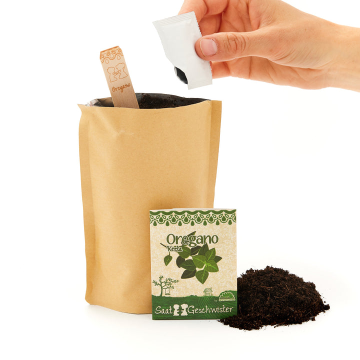 Hand hält ein Päckchen mit Oregano-Samen neben einer Papiertüte mit Pflanzerde und einem Holzetikett mit Oregano-Aufschrift.