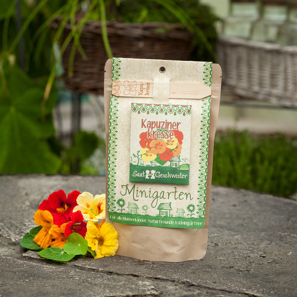 Verpackung für Kapuzinerkresse Samen mit dem Titel "Minigarten" neben blühenden Kapuzinerkresse-Blüten auf einer Steinfläche.