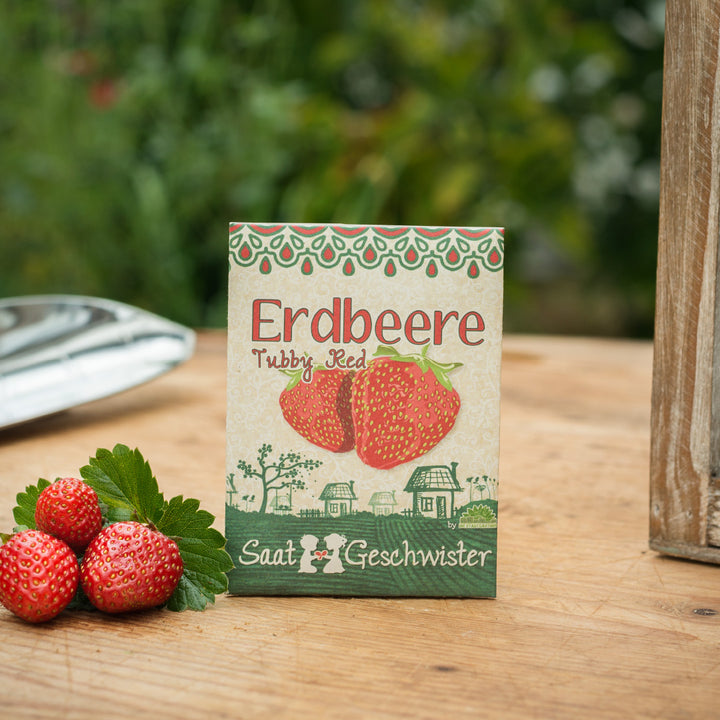 Erdbeersamentütchen mit der Aufschrift "Erdbeere Tubby Red" und echten Erdbeeren im Vordergrund auf einem Holztisch.