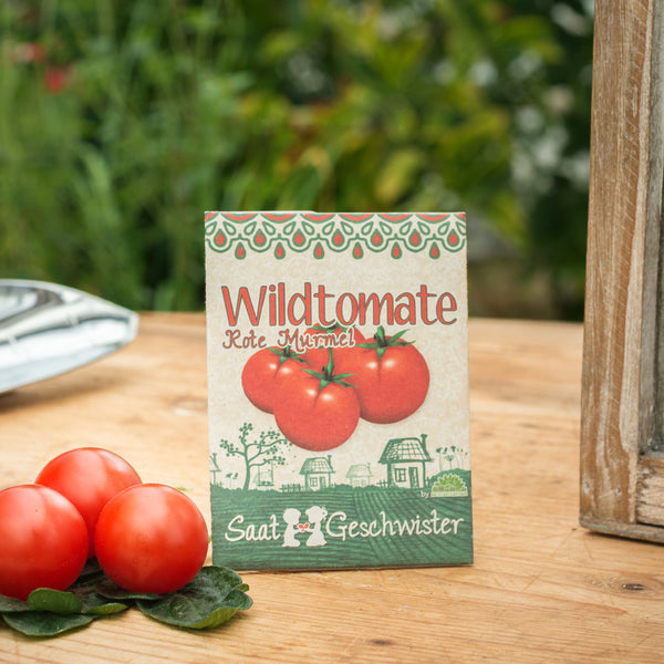 Päckchen mit Saatgut für Wildtomaten neben frischen Tomaten auf einer Holzoberfläche mit Gartenhintergrund