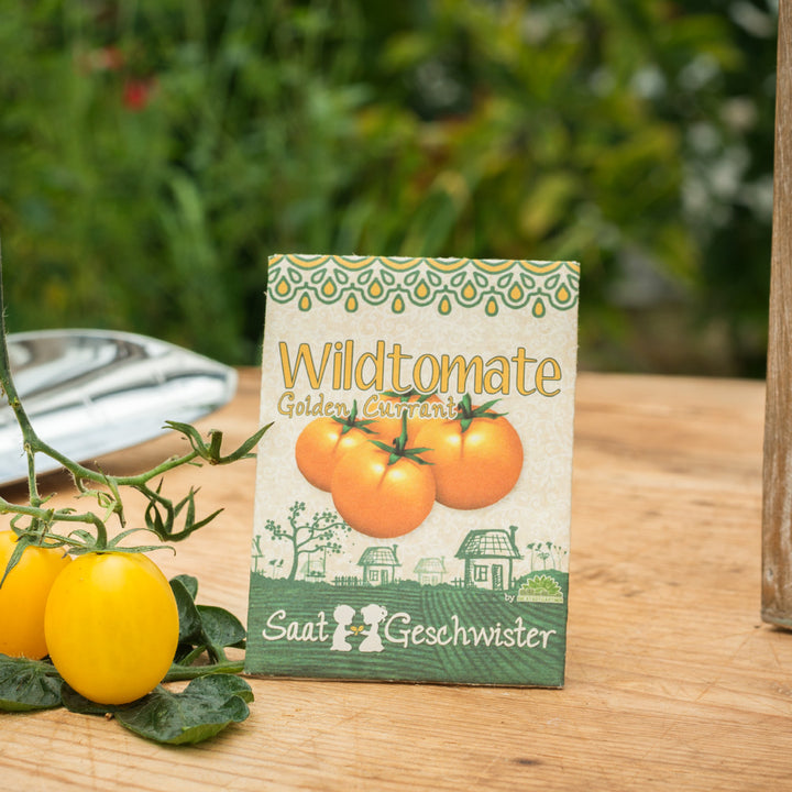 Päckchen mit Samen von Wildtomaten "Golden Currant" auf einem Holztisch mit reifen gelben Tomaten im Vordergrund und unscharfem Grünpflanzenhintergrund.