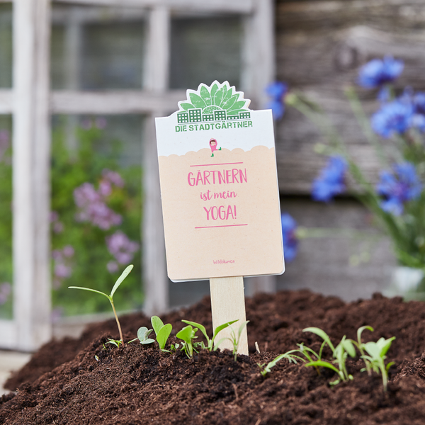 Schild im Garten mit der Aufschrift "Gärtnern ist mein Yoga!" steht in frisch gepflanzter Erde.