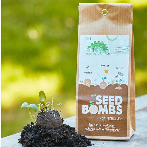 "Verpackung von 'Seed Bombs' mit Keimlingen auf Erde im Vordergrund und unscharfem grünen Hintergrund"