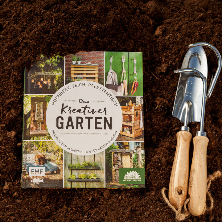 Gartenbuch mit dem Titel "Dein kreativer Garten" auf Erdboden neben Gartengeräten.
