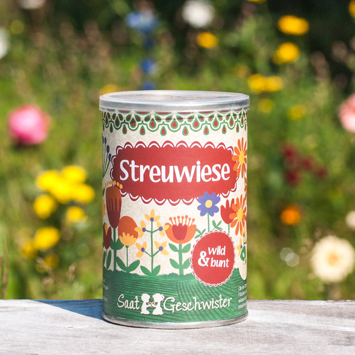 Farbenfrohe Saatgutdose mit der Aufschrift "Streuwiese" vor unscharfem Blumenhintergrund.